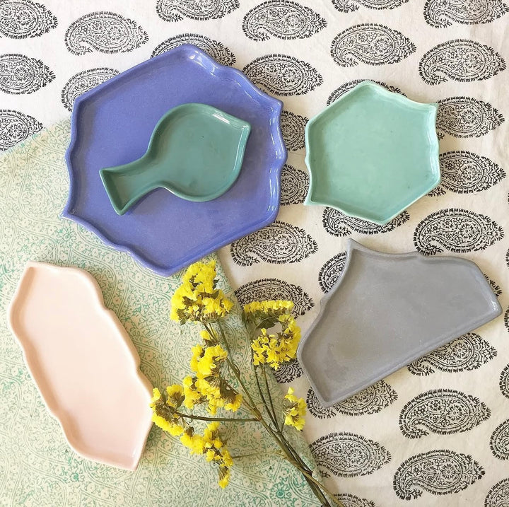 Four irregular ceramic dish pieces in pastel colors arranged on a patterned fabric with yellow flowers. The set includes a lavender dish holding a mint green leaf-shaped piece, a mint green hexagonal dish, a pale pink elongated dish, and a gray organic-shaped dish.