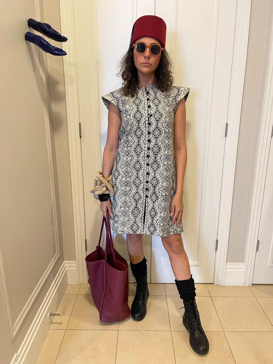 A woman stands in a hallway wearing a sleeveless black and white patterned dress, a red cap, sunglasses, black boots, and holding a burgundy tote bag.