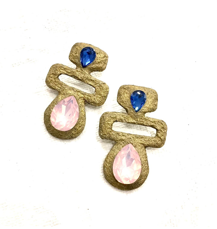A pair of sculptural statement earrings with a textured gold-tone metal frame, featuring a small faceted blue teardrop gemstone at the top and a larger pearlescent light pink teardrop gemstone at the bottom, shown on a plain white background.