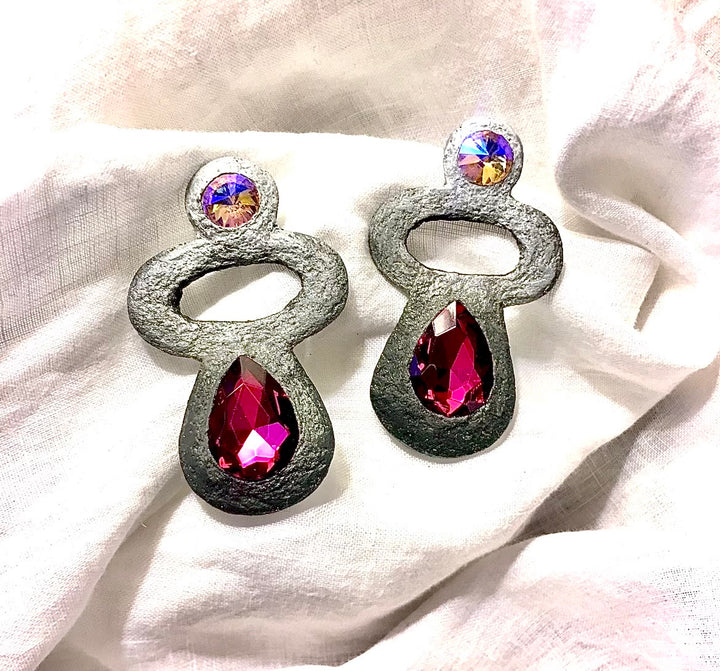 A pair of silver-toned statement earrings with teardrop-shaped magenta crystal drops and small iridescent round crystals at the top, displayed on a soft white fabric background.