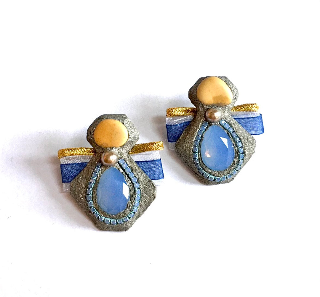 A pair of vintage-style angel earrings with blue teardrop-shaped stones, textured gray stone bodies, gold accents, and blue and white ribbon wings, shown on a plain white background.