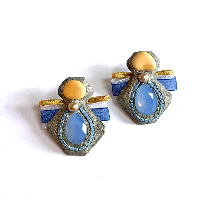 A pair of vintage-style angel earrings with blue teardrop-shaped stones, textured gray stone bodies, gold accents, and blue and white ribbon wings, shown on a plain white background.