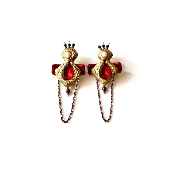 A pair of ornate gold-toned earrings with red gemstone-like centers, small white pearl accents, dark bead details, and hanging chain drops, shown on a plain white background.