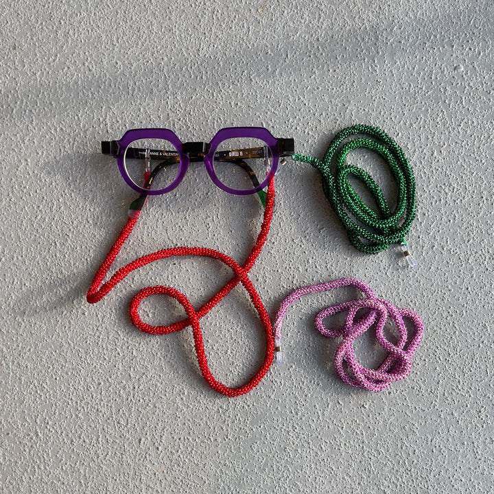 A pair of eyeglasses with a purple frame and black temples, each attached to a thick braided rope strap in a different color: red, green, and pink. The glasses and rope set are arranged on a textured light gray surface.