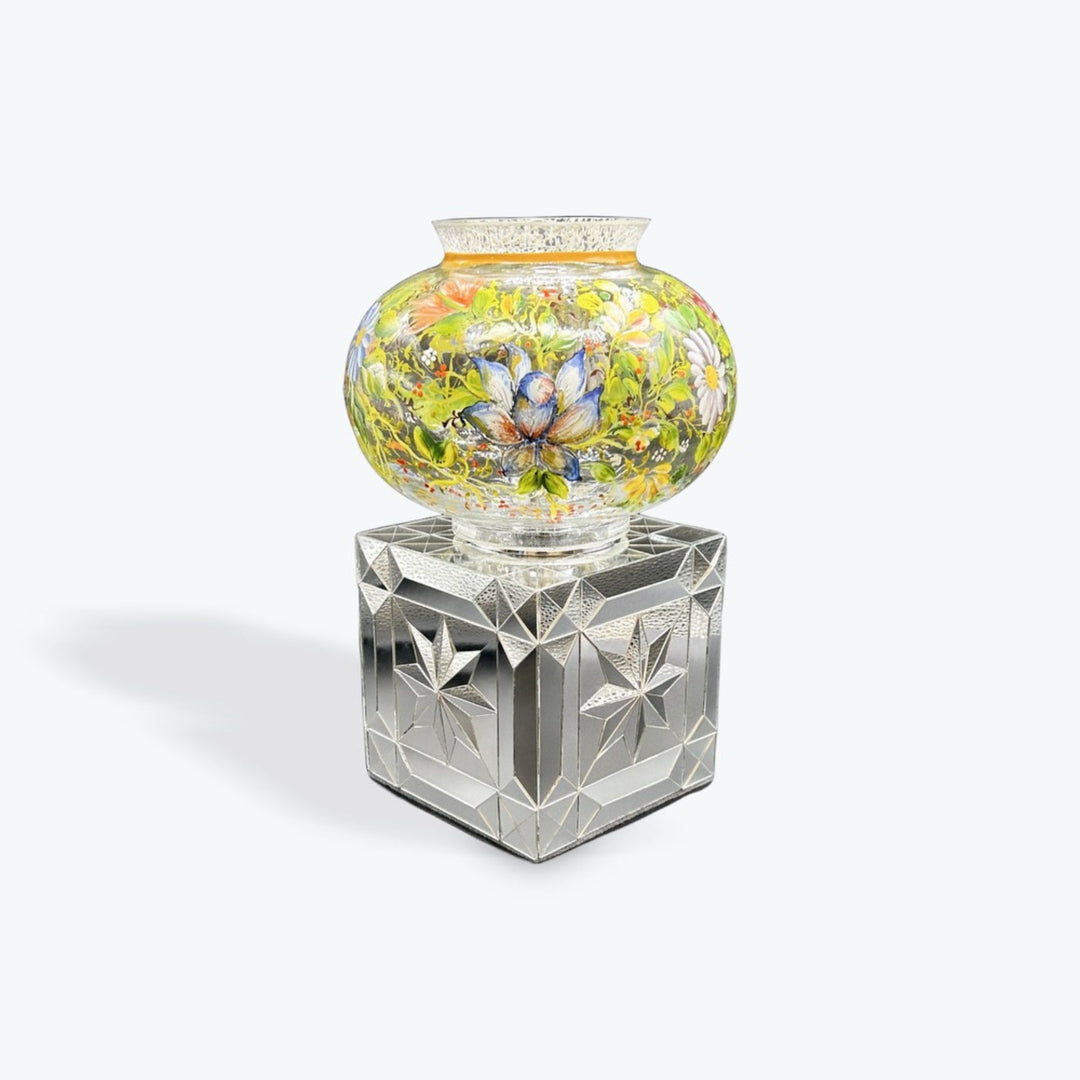 A decorative glass vase with a colorful floral pattern, displayed on a faceted clear glass pedestal against a light gray background.